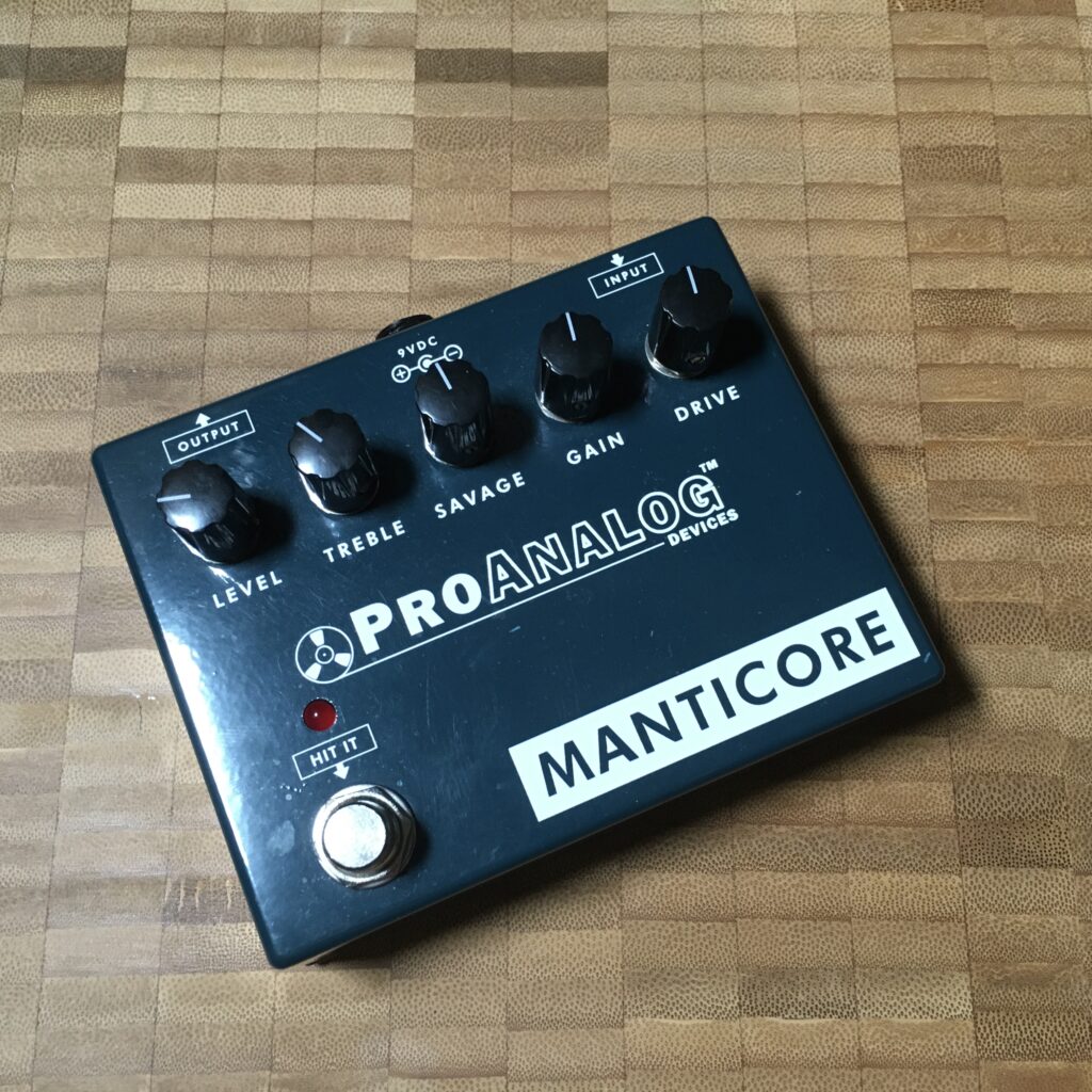 This photo shows a ProAnalog Devices Manticore v2 pedal against a butcher's block wooden background. The pedal is finished in a very hard to photograph dark green.

The pedal's five controls are clearly visible: Level, Treble, Savage, Gain and Drive. Also visible is the single on/off stomp switch.