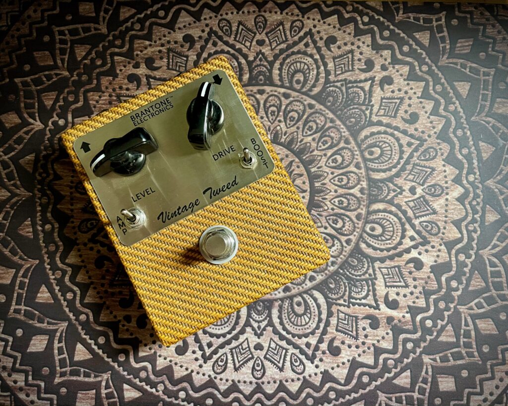 This photo shows a Vintage Tweed pedal by Brantone Electronics. The pedal itself is noticeable because it is covered in the same striped yellow cloth that was used on vintage tweed amps.