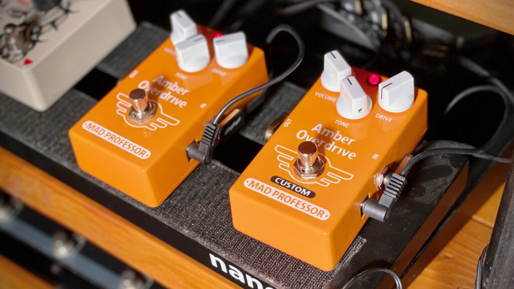 This photo is a close-up of my pedal board.

There are two pedals shown: the original Amber Drive, and the factory-modded Amber Drive with Midas Touch Mod.

The only visual way to tell the two pedals apart is the black 'CUSTOM' sticker that has been added below the footswitch on the modded pedal.

Apart from that, the two pedals look identical.

Both have 'Volume', 'Drive' and 'Tone' controls on the top, along with side jacks and a power jack next to the 'In' jack on the right-hand side of the pedal.