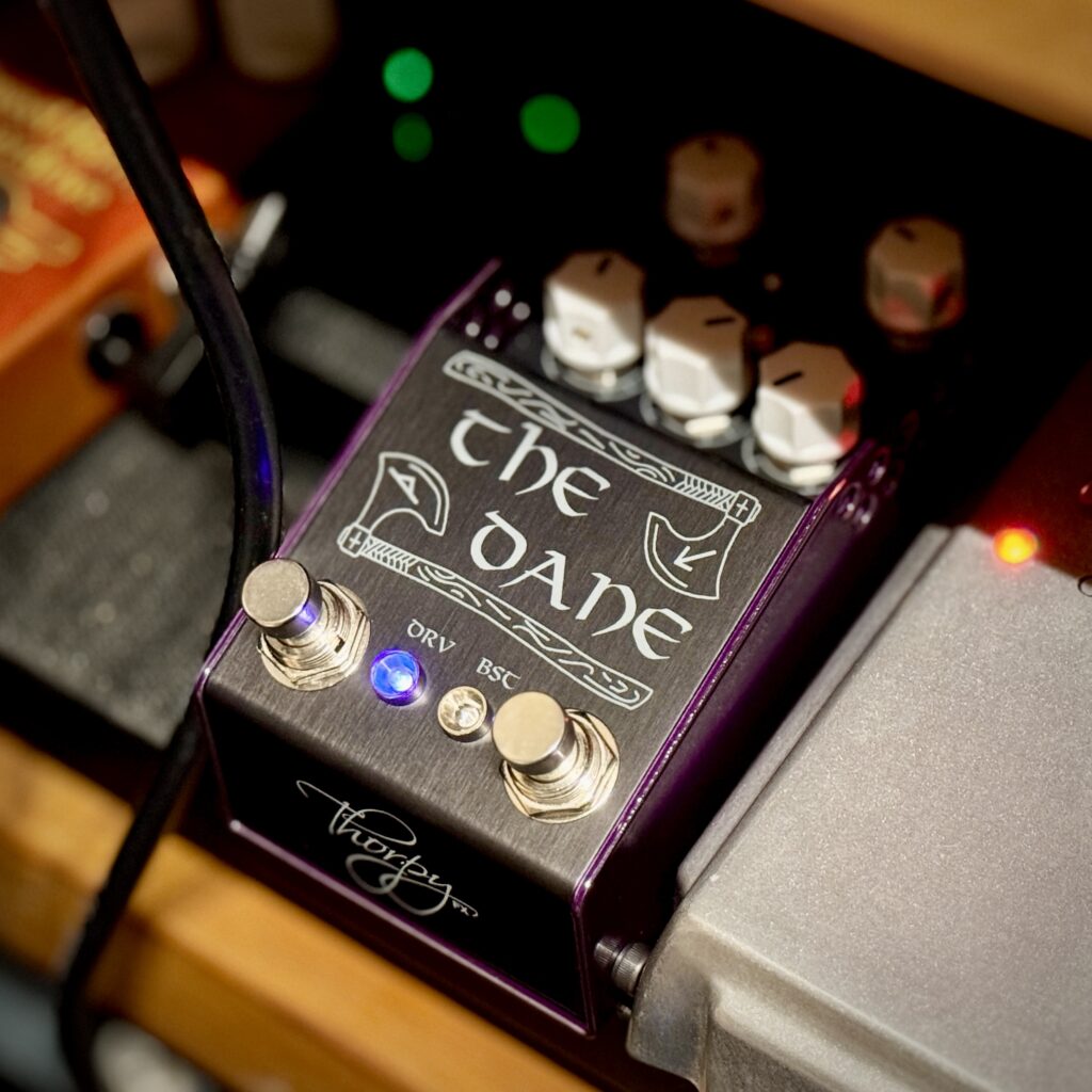 The Dane mk2 guitar pedal from ThorpyFX. A brushed black gunmetal top, with five white control knobs and two foot switches. Purple accent sides.