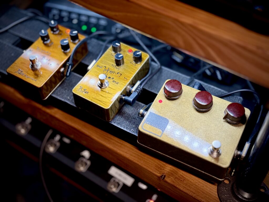 This is a photo of three guitar effects pedals on the author's pedalboard.
On the far-left, is the BJFe-in-your-hand series Honey Bee Overdrive by One Control. It's a distinctive warm honey colour, with four control knobs on the top surface.
In the middle is the BJFe Honey Bee OD Retro, by BJFe. This pedal is a lighter shade, almost yellow. It has three control knobs on its top surface.
On the right-hand side is the Klone v2 by Arc Effects. This pedal has a metallic gold finish. It spots the Klon's distinctive control knobs.