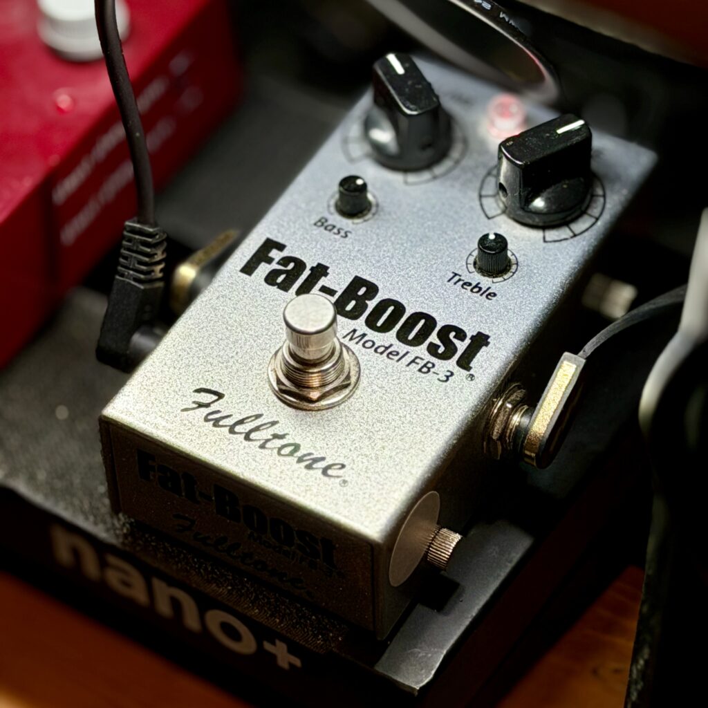 This is a photo of my Fulltone Fat-Boost FB-3 guitar effects pedal.
The pedal comes in a metallic silver case. Input and output audio jacks are on the side. 9V power is on the left-hand side of the pedal, close to the output audio jack.
There are five controls shown on the top of the pedal:
- two large knobs at the top, for output volume and drive (a bias control for the internal FET)
- two small trim pots, one labelled "Bass" and one labelled "Treble"